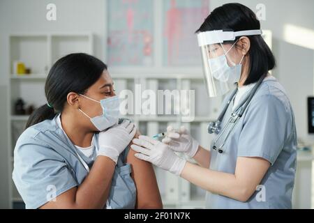 Young Asian nurse in protective workwear making injection to her mixed-race female colleague during vaccination against covid in clinics Stock Photo
