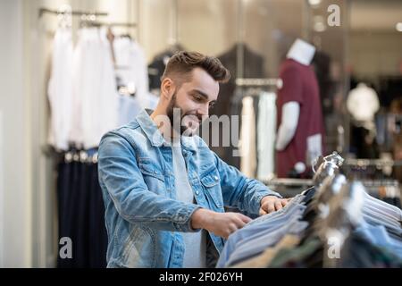 Young bearded man choosing new clothes on the rack in shopping mall Stock Photo