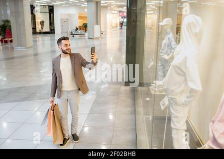Young man making photo of mannequin on the phone behind the window while doing shopping in the mall Stock Photo