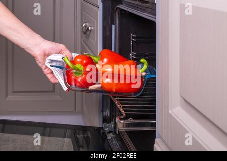 Woman housewife places a fireproof glass tray of ripe red bell peppers for baking in an electric oven. Stock Photo