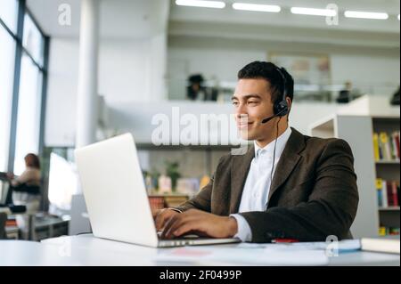 Smiling manager at the workplace, uses laptop. Handsome male hispanic employee works on project or new startup, sit at the desk in office, wearing headset, communicate with colleagues Stock Photo