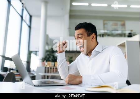 Excited businessman happy about work results, feeling euphoric. Overjoyed male freelancer or employee sitting at the desk, well done with project or got a good news Stock Photo