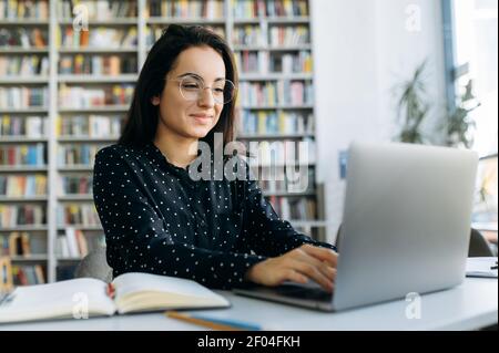 Attractive female programmer at the workplace. Pretty woman using laptop, developing a new project or app, sit at the table in modern office, smiles Stock Photo