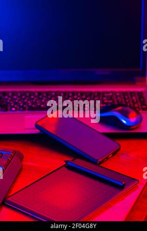 Digital designer workplace. Remote work. Freelancer workplace in neon light. Computer, laptop, graphics tablet and smartphone on the table Stock Photo