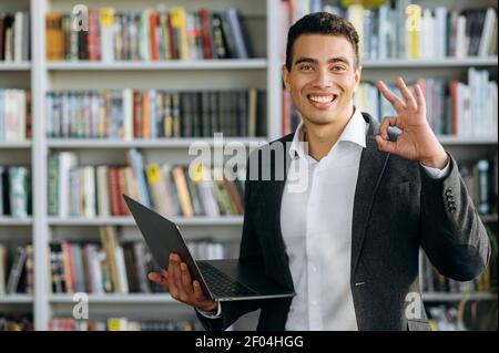 Hispanic influential young businessman in suit is standing in office, posing, showing okay sign. Successful male entrepreneur stands with laptop, looks at the camera, smiling Stock Photo