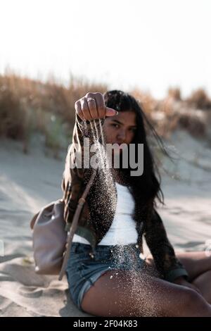 A shallow focus of a cool young Hispanic female sitting on a beach and throwing sand on it Stock Photo