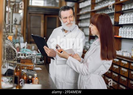 Confident female pharmacist talking with her senior male colleague about side effects of a medicine or pharmaceutical product for sale, working in old Stock Photo
