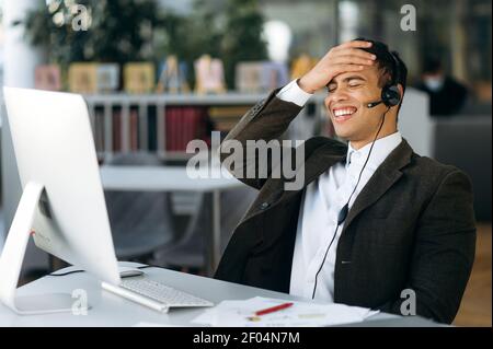 Cheerful businessman or freelancer in headset, smiles during conversation by video call. Joyful male manager or consultant sit at the work desk in modern office uses computer Stock Photo