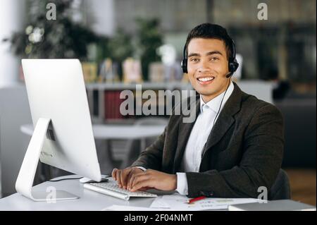 Portrait of male employee of call center. Smiling hispanic man looks at the camera, sitting in modern office wearing headset, conducts video meeting, support service concept Stock Photo