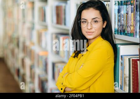 Portrait of elegant young pretty woman. Beautiful female teacher, freelancer or student looks at the camera, smiling. Young stylish lady wearing eyeglasses holds in arms some books, standing in Stock Photo
