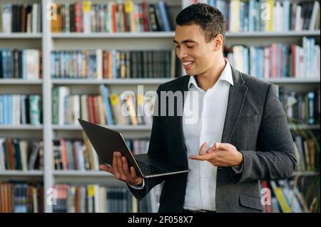 Successful male employee is using laptop, standing in office or library, searching some information for project. Happy hispanic businessman looking at the laptop screen, smiling Stock Photo