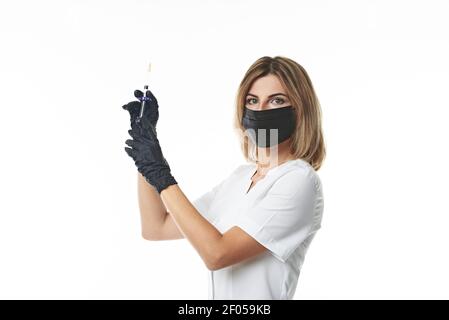 Side portrait isolated on white background of a young female doctor in a medical protective mask and surgical gloves holding a syringe with a medicati Stock Photo