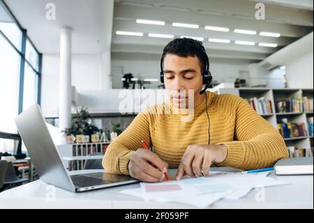 Concentrated hispanic successful freelancer or student sitting at his work desk, makes notes, uses laptop for remote work or distance learning Stock Photo