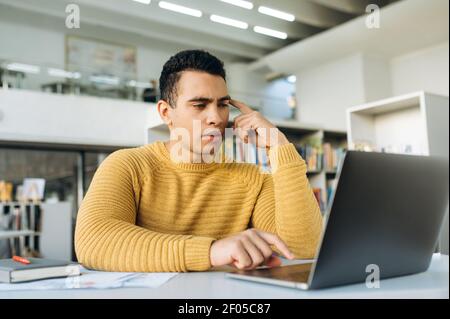 Serious hispanic guy sit at the work desk, using laptop. Concentrated male employee looking at the screen, browsing internet, search ideas, working on project Stock Photo