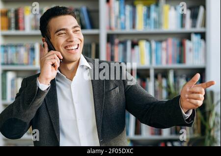 Cheerful employee using smartphone, having a conversation. Happy businessman standing in modern office, smiling, talks with friends or colleagues while working Stock Photo