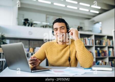 Happy hispanic male consultant sit at the desk, looks at the camera and smiles. Attractive young man in headphones communicate with clients, work in support service Stock Photo