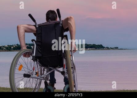 Young man is sitting on wheelchair near sea at sunset. Stock Photo