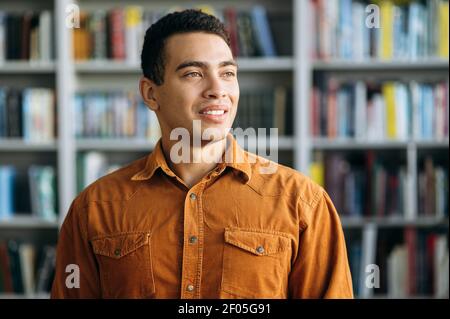 Portrait of a satisfied male freelancer or student. Confident successful hispanic business man looks away and smiling, standing in modern office or university library Stock Photo