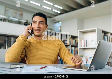 Cheerful hispanic man uses cell phone, talking, smiling. Happy male employee sit at the desk, talking with friends or colleagues while working, communication concept Stock Photo