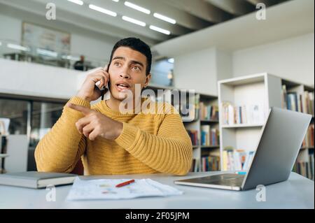Successful confident male freelancer, talks with colleague by cell phone. Hispanic business man sit at the table in modern office, using smartphone and laptop Stock Photo