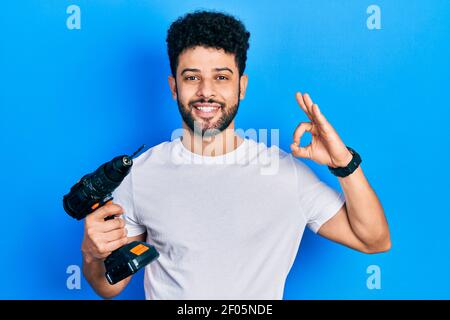 Young arab man with beard using drill doing ok sign with fingers, smiling friendly gesturing excellent symbol Stock Photo