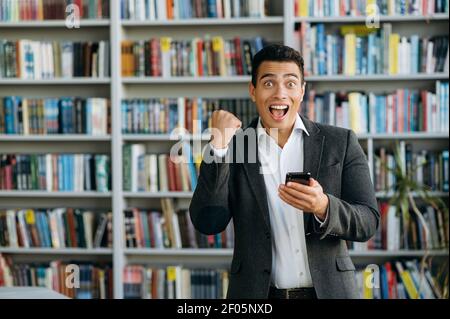 Excited hispanic male manager standing in office, uses smartphone, smiling. Happy male freelancer or student got a pleasant news, wins a lottery, success concept Stock Photo