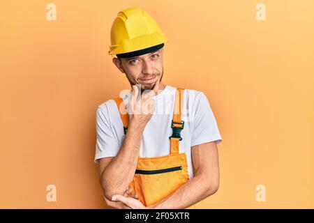 Hispanic young man wearing builder uniform and safety hardhat thinking concentrated about doubt with finger on chin and looking up wondering Stock Photo