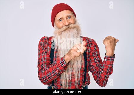 Old senior man with grey hair and long beard wearing hipster look with wool cap pointing to the back behind with hand and thumbs up, smiling confident Stock Photo