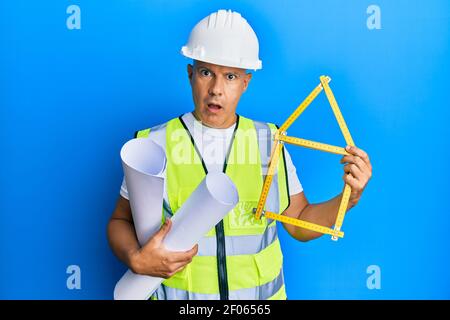 Middle age bald man wearing architect hardhat holding build project in shock face, looking skeptical and sarcastic, surprised with open mouth Stock Photo