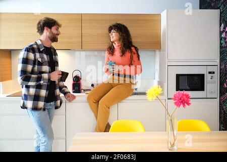Young cheerful couple in bright casual clothes talking in cosy kitchen and looking at each other Stock Photo