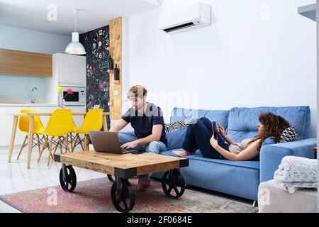 Young Woman browsing tablet lying on sofa while man surfing internet on laptop in modern apartment Stock Photo