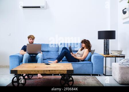 Young Woman browsing tablet lying on sofa while man surfing internet on laptop in modern apartment Stock Photo