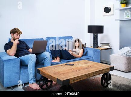 Young Woman browsing tablet lying on sofa while man surfing internet on laptop in modern apartment Stock Photo