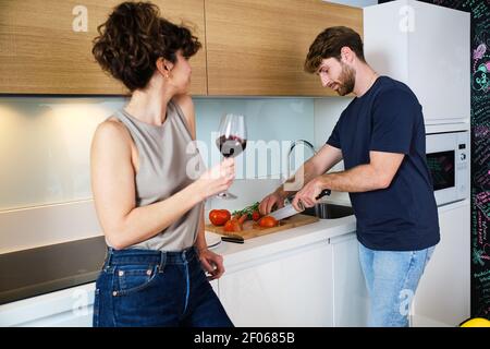 Side view of young couple standing in kitchen with glass of red wine near stove and counter and cutting tomatoes on cutting board with knife near sink Stock Photo