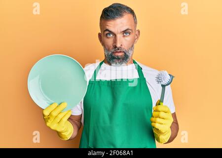 Middle age handsome man wearing apron holding scourer washing dishes in shock face, looking skeptical and sarcastic, surprised with open mouth Stock Photo