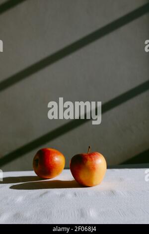 Fresh whole apples placed on white tablecloth against gray wall with shadows in daylight Stock Photo