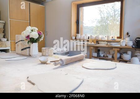 Table with smoothed pieces of clay near rolling pin and bowl near vase with flowers near window with  handmade pots on windowsill and plates on table Stock Photo
