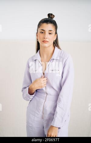 Unemotional young thoughtful female in stylish overall looking at camera standing in studio background Stock Photo