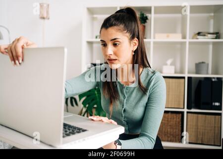 Young attentive remote employee opening modern portable computer against shelves at home in daylight Stock Photo