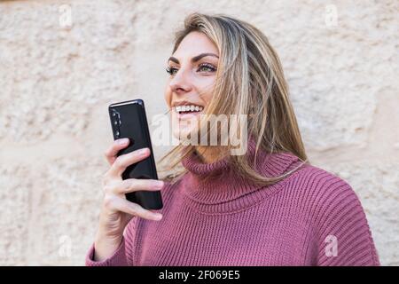 Tender charming female standing in city street with smartphone recording a voice near stone old wall looking away Stock Photo