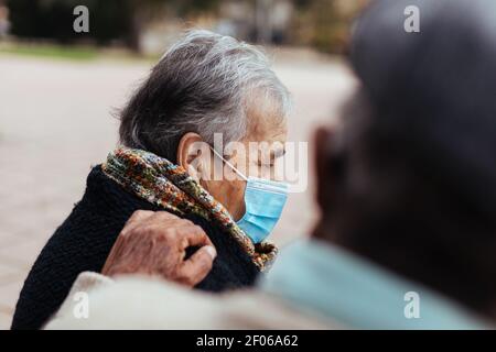 Side view of elderly couple sitting on a park bench while the husband puts his hand on his wife's shoulder in a gesture of love Stock Photo