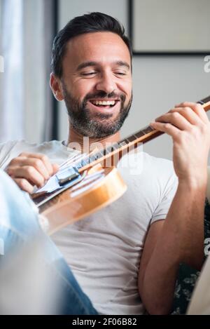 Happy adult ethnic male musician in casual clothes relaxing on comfortable sofa and playing acoustic guitar at home Stock Photo