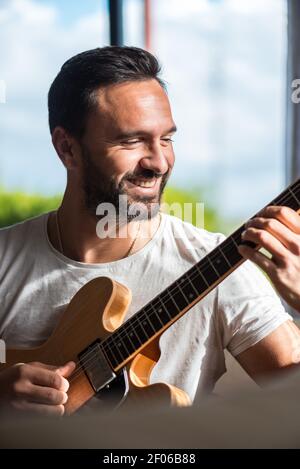 Happy adult ethnic male musician in casual clothes relaxing on comfortable sofa and playing acoustic guitar at home Stock Photo