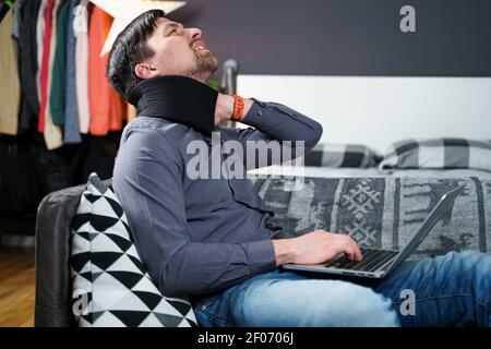 Man employee after accident working at home behind laptop and feels pain in neck, uses black neck collar support. Male with neck injury freelance Stock Photo