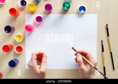 Hands holding paintbrush ready to pain with colored paints on blank paper at the table. Art and creativity concept. Stock Photo