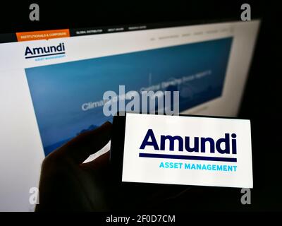 Person holding smartphone with logo of French investment company Amundi S.A. on screen in front of website. Focus on phone display. Unmodified photo. Stock Photo