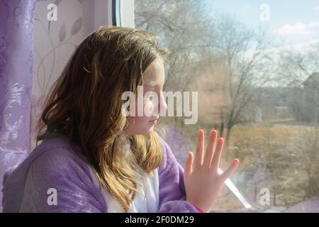 Teenage girl looks out window at dawn in sun. Hand on glass. Close-up, side view, selective focus. Stock Photo