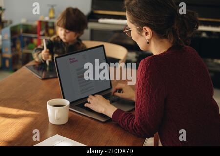 Woman working on laptop with her son using digital tablet in background. Working mother with son playing with tablet computer. Stock Photo