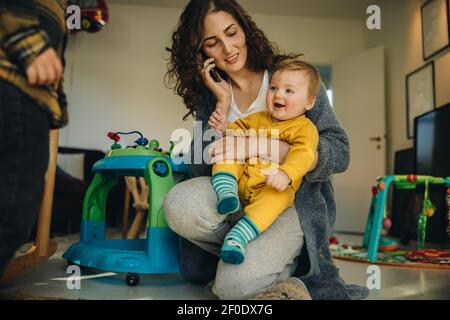 Woman talking on mobile phone while taking care of a his son at home. Mother holding baby while talking on phone. Stock Photo