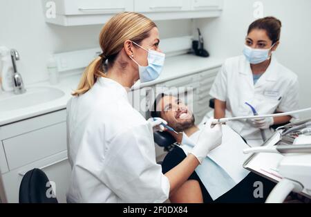 Dental doctor looking at a display monitor while treating a male patient. Female dentist wearing face mask with male patient sitting in dentist's chai Stock Photo